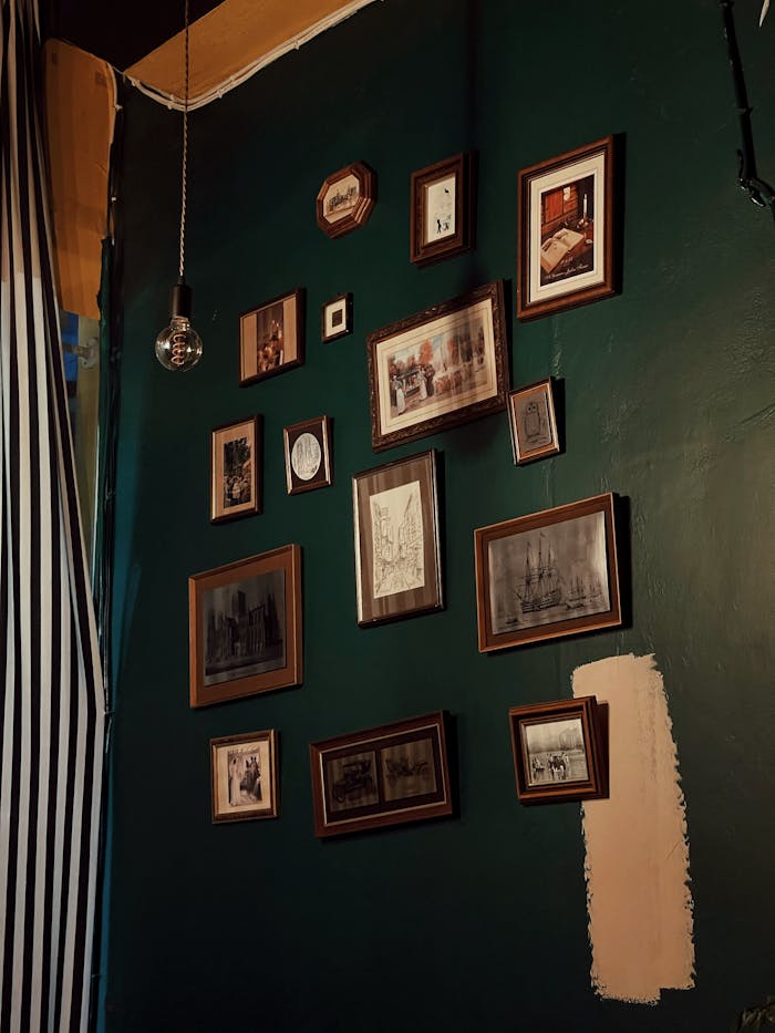 A collection of vintage picture frames arranged on a dark green wall with a hanging light bulb.